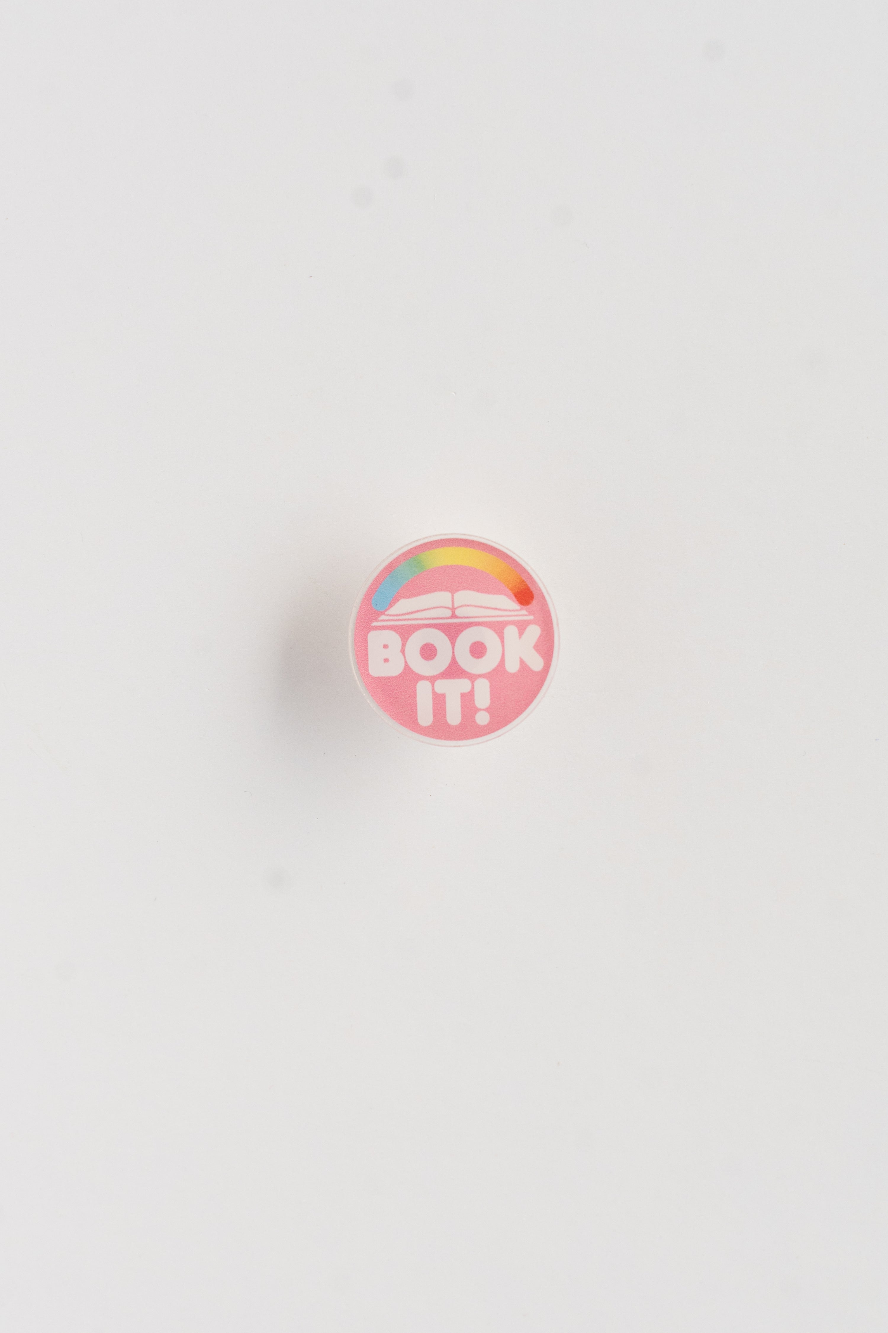 Round pink button with 'BOOK IT!' text and rainbow design on a white background