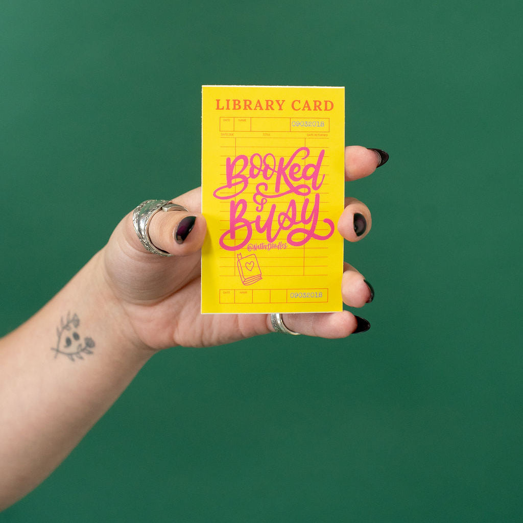 Hand holding a yellow 'Booked & Busy' library card against a green background