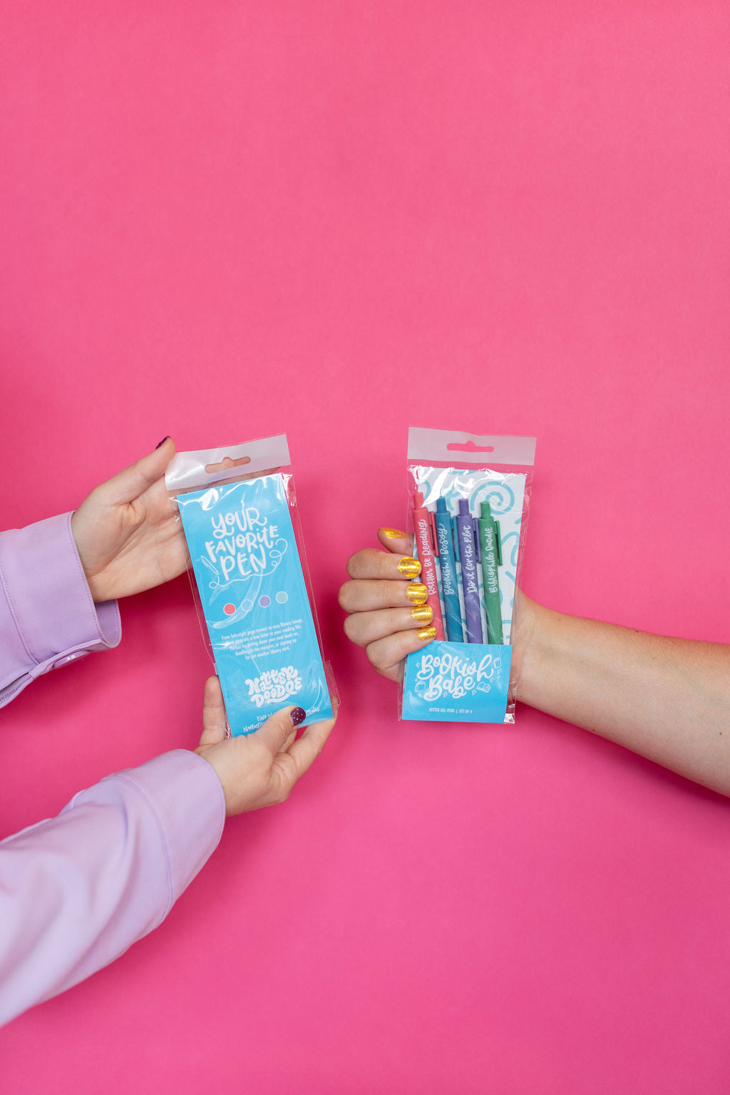 Two hands holding packages of colorful pens against a pink background