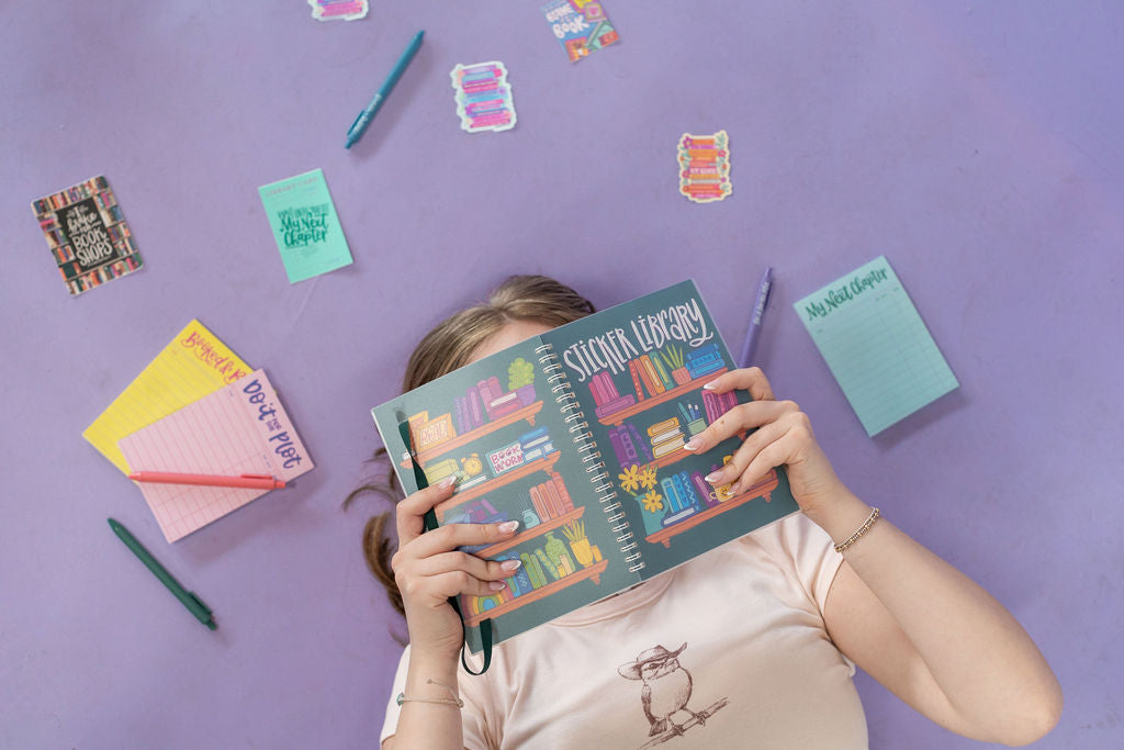 Person holding a colorful notebook with stickers and stationery items on a purple background