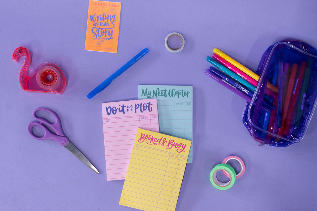 Assorted stationery items including pens, scissors, tape, and notepads on a purple background