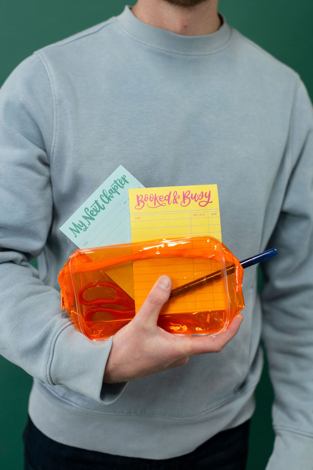 Person holding an orange pencil pouch with a yellow and blue notepad in front of a green background