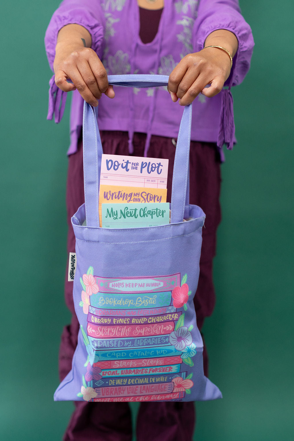 Person holding a purple tote bag with book-related text and graphics against a green background