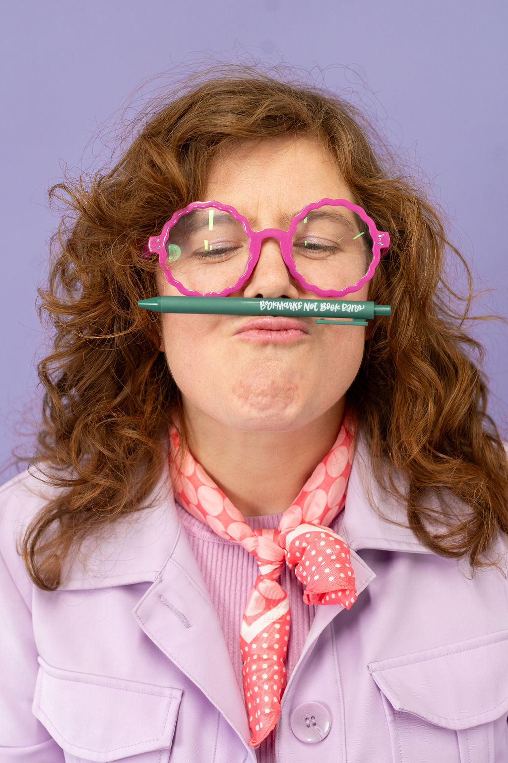 Person with pink glasses holding a green pencil between their lips against a purple background