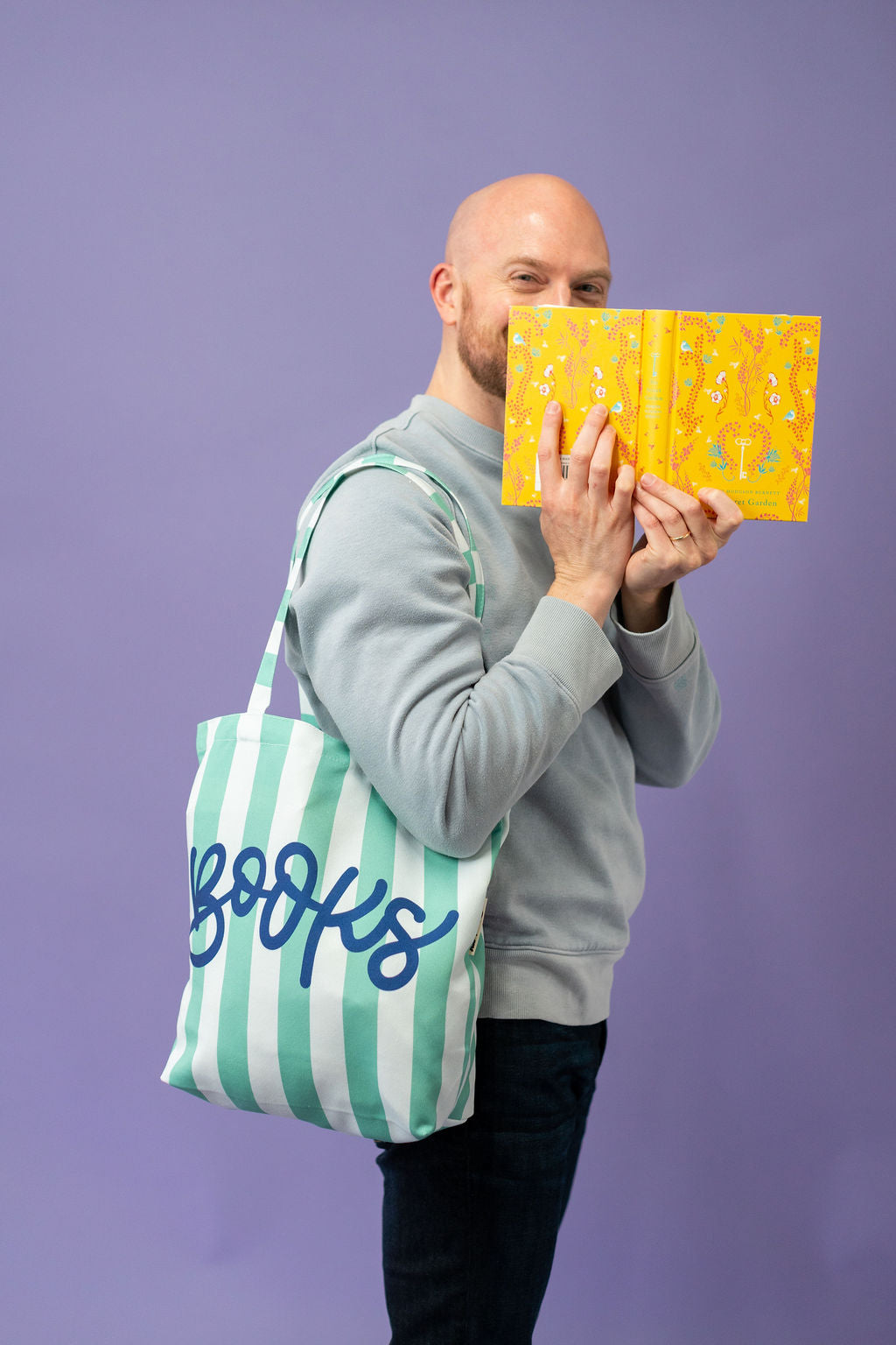 Man holding a yellow book with a green and white striped bag labeled 'books' on a purple background