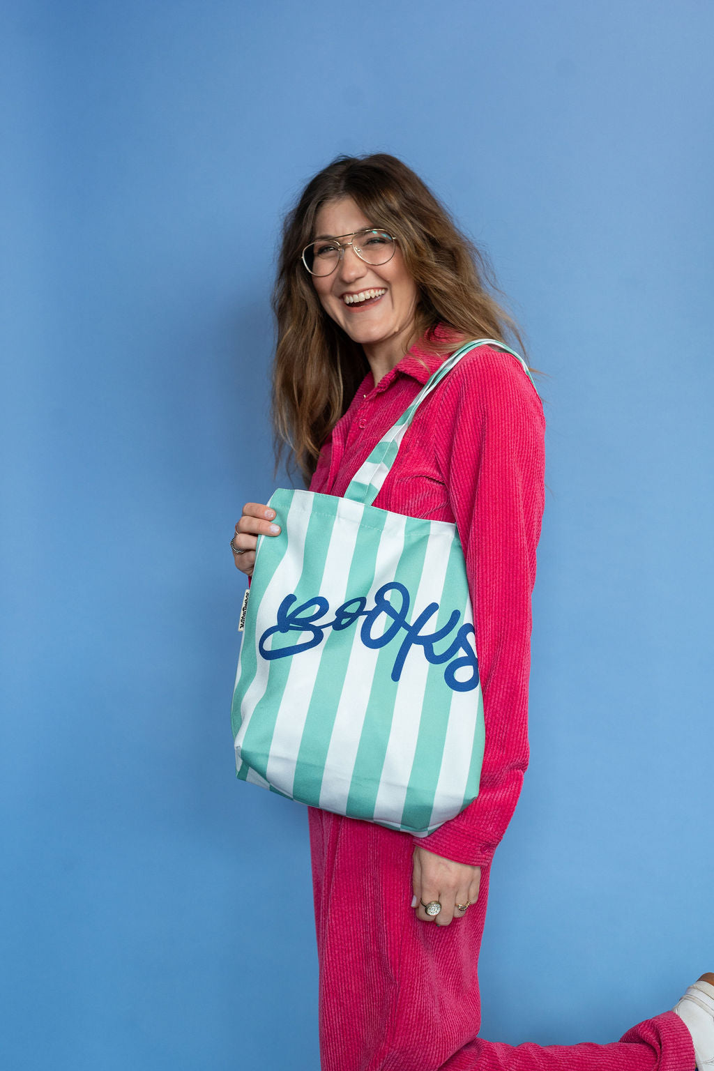 Woman holding a green and white striped tote bag with 'Books' written on it against a blue background