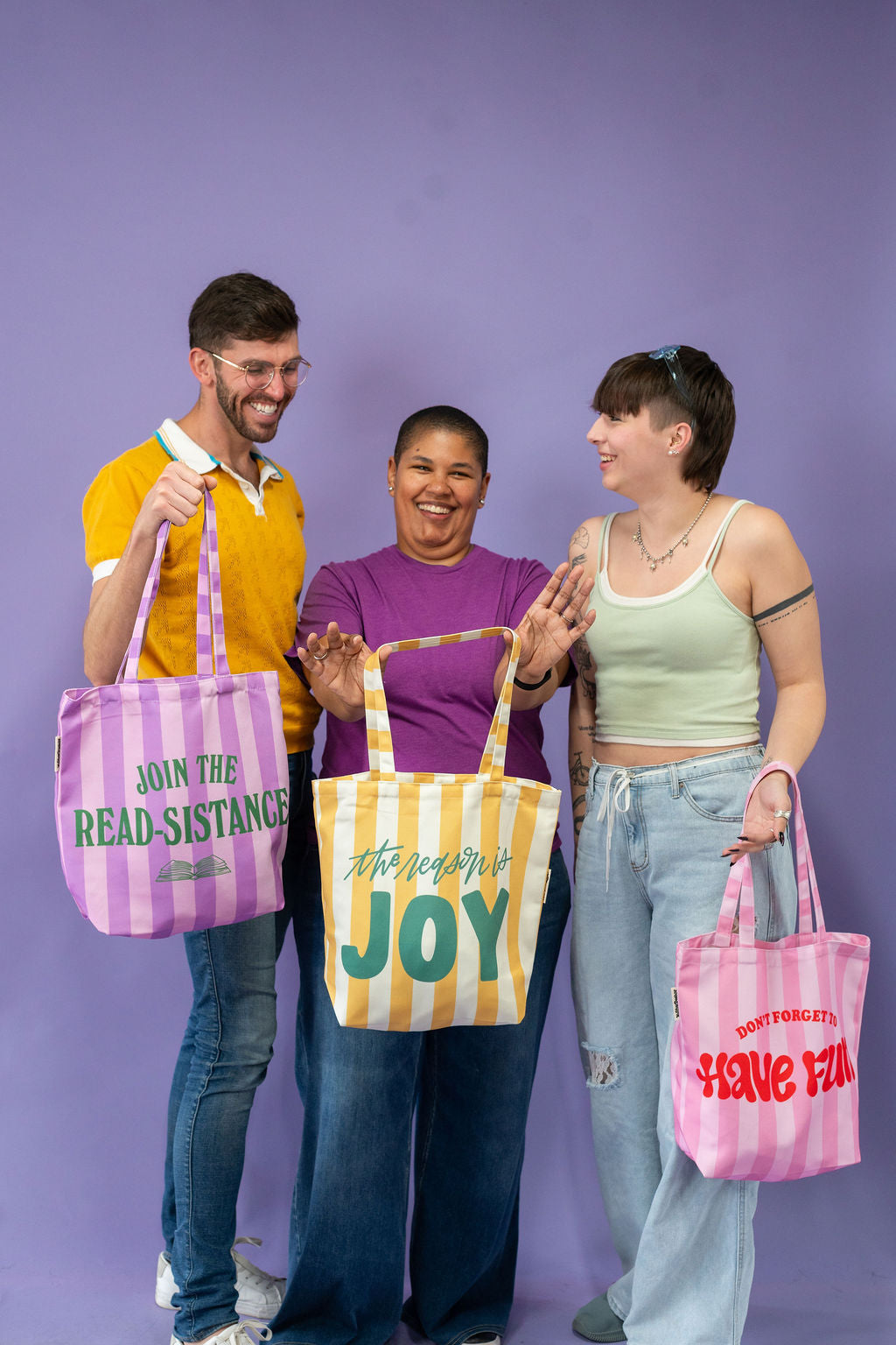 Three people holding colorful tote bags with text against a purple background