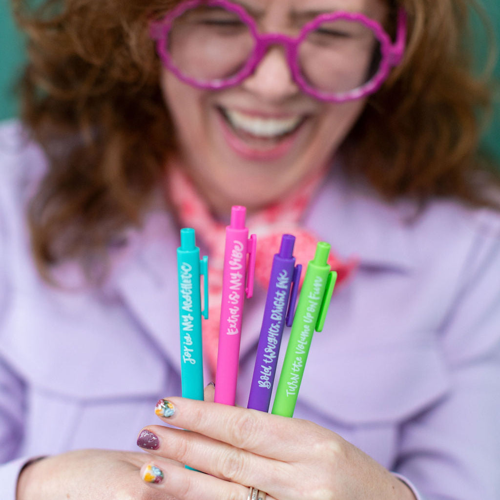 Person wearing large pink glasses holding colorful pens with visible text.