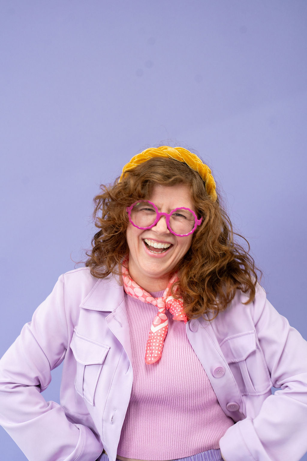 Person wearing a pink jacket, pink sweater, yellow headband, and pink glasses against a purple background