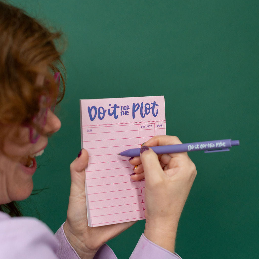 Person holding a pink notepad titled 'Do It For The Plot' with a pen against a green background