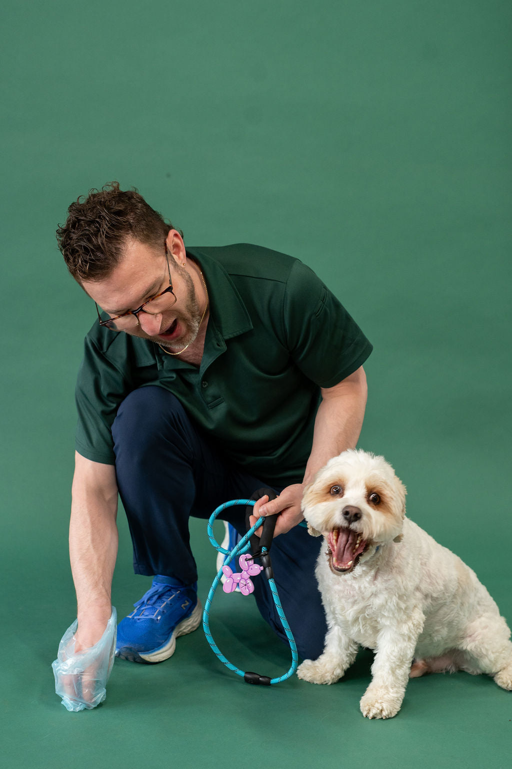 Man in green shirt and blue pants kneeling with a small white dog on a green background