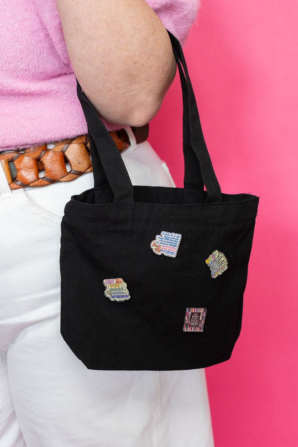 Black tote bag with colorful pins held by a person against a pink background
