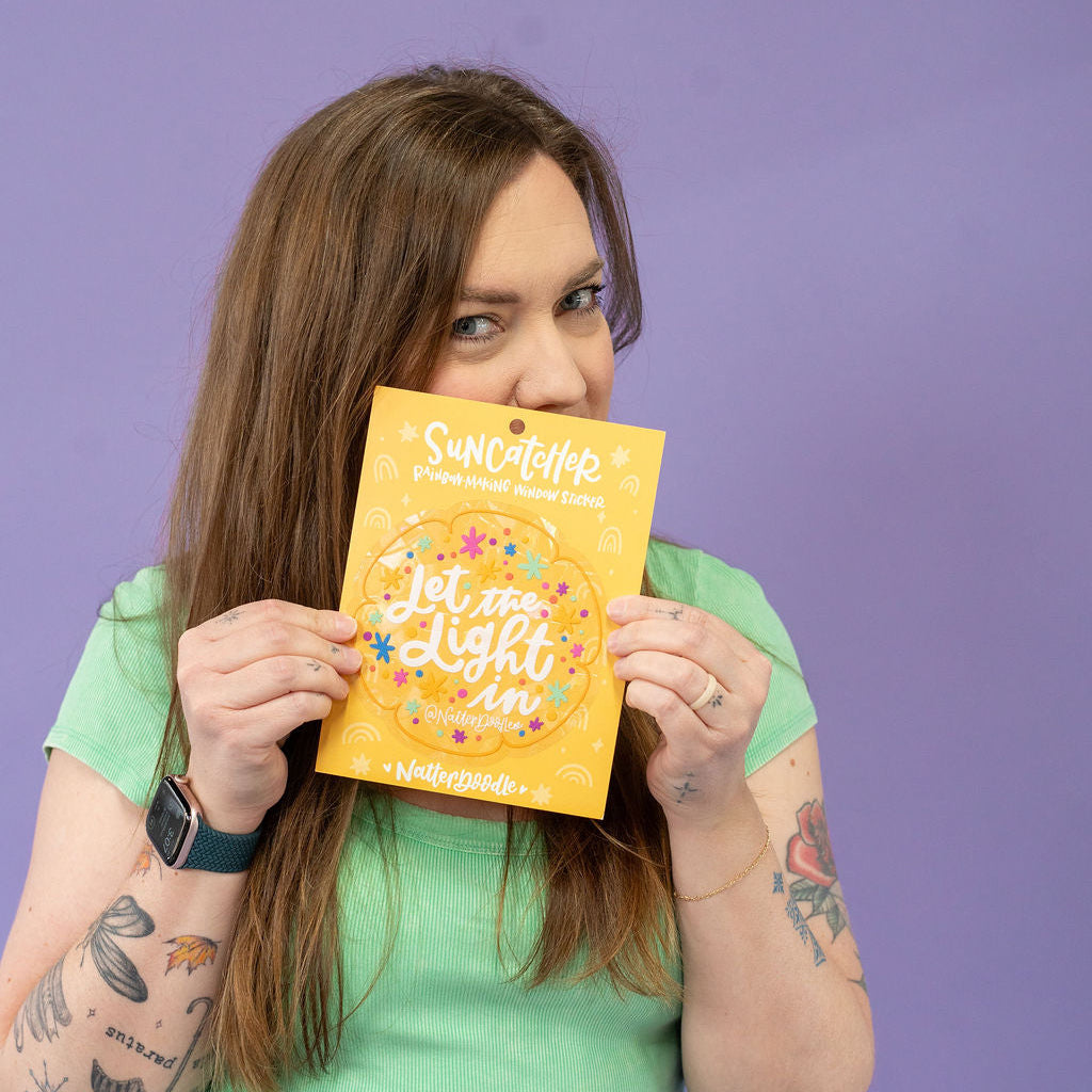 Person holding a yellow suncatcher titled 'Let the Light In' against a purple background