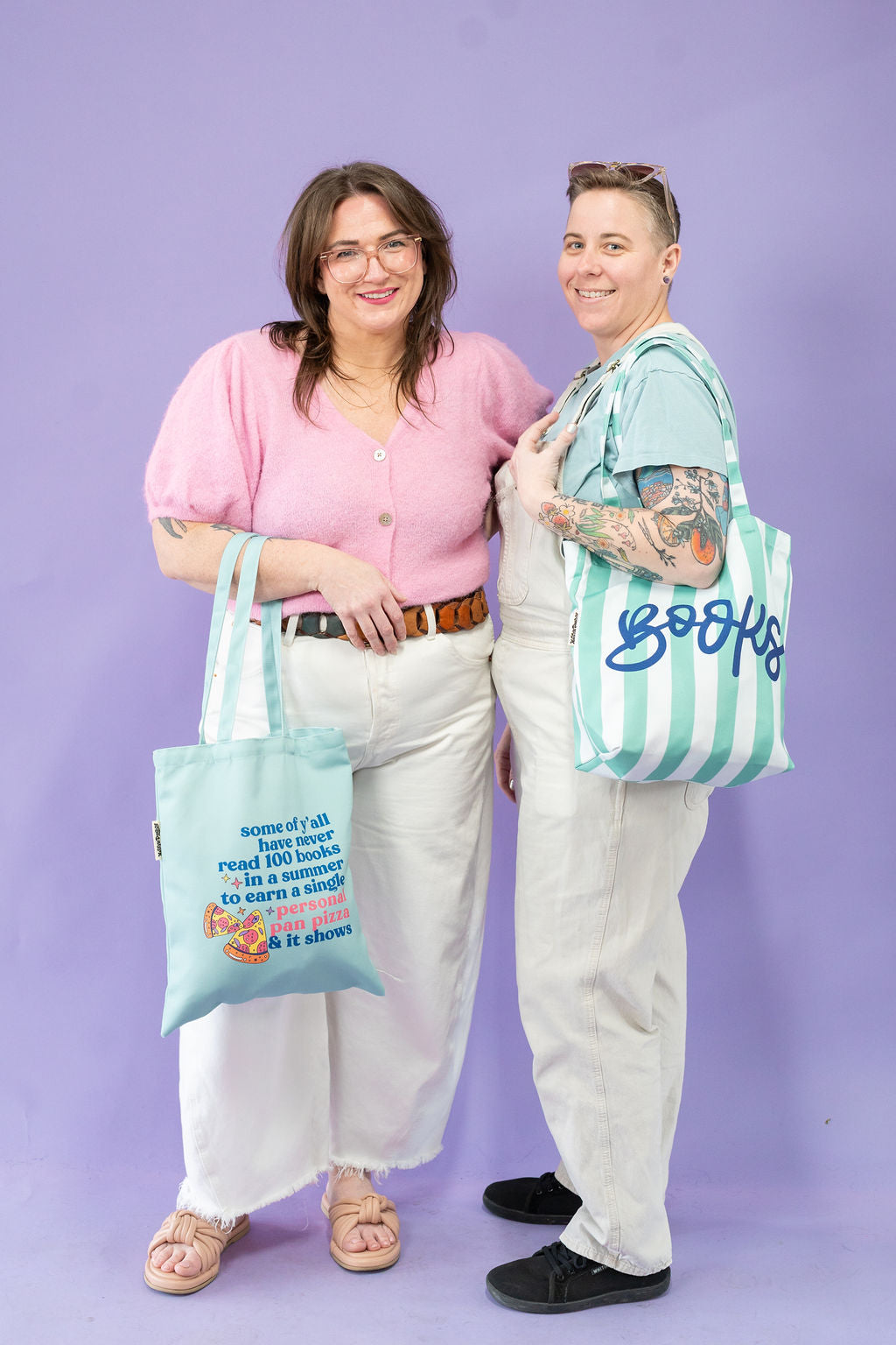 Two people standing side by side against a purple background, each holding a tote bag.