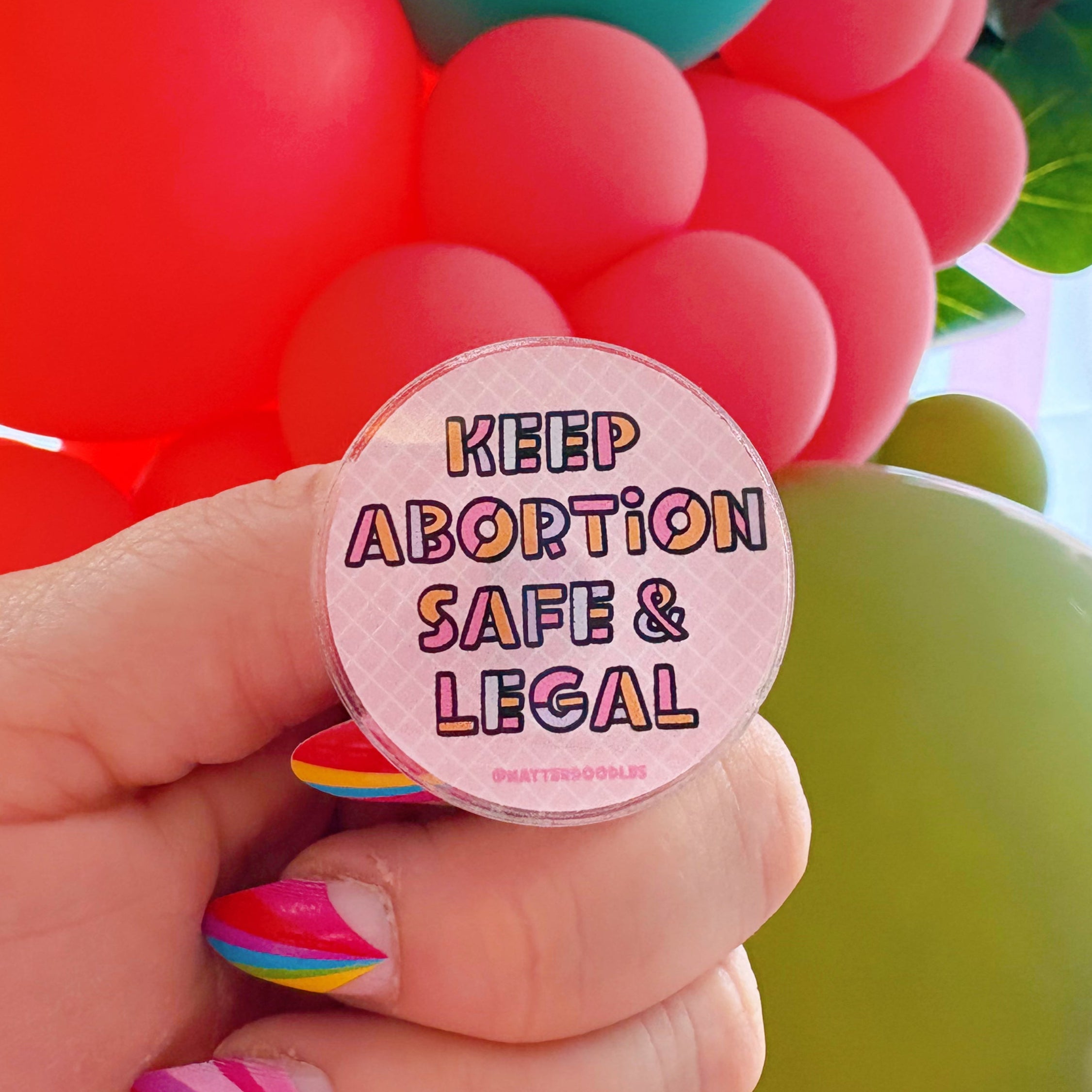 A hand holding a round acrylic pin with the text 'KEEP ABORTION SAFE & LEGAL' in bold letters, against a pastel pink grid background with multicolored edges.