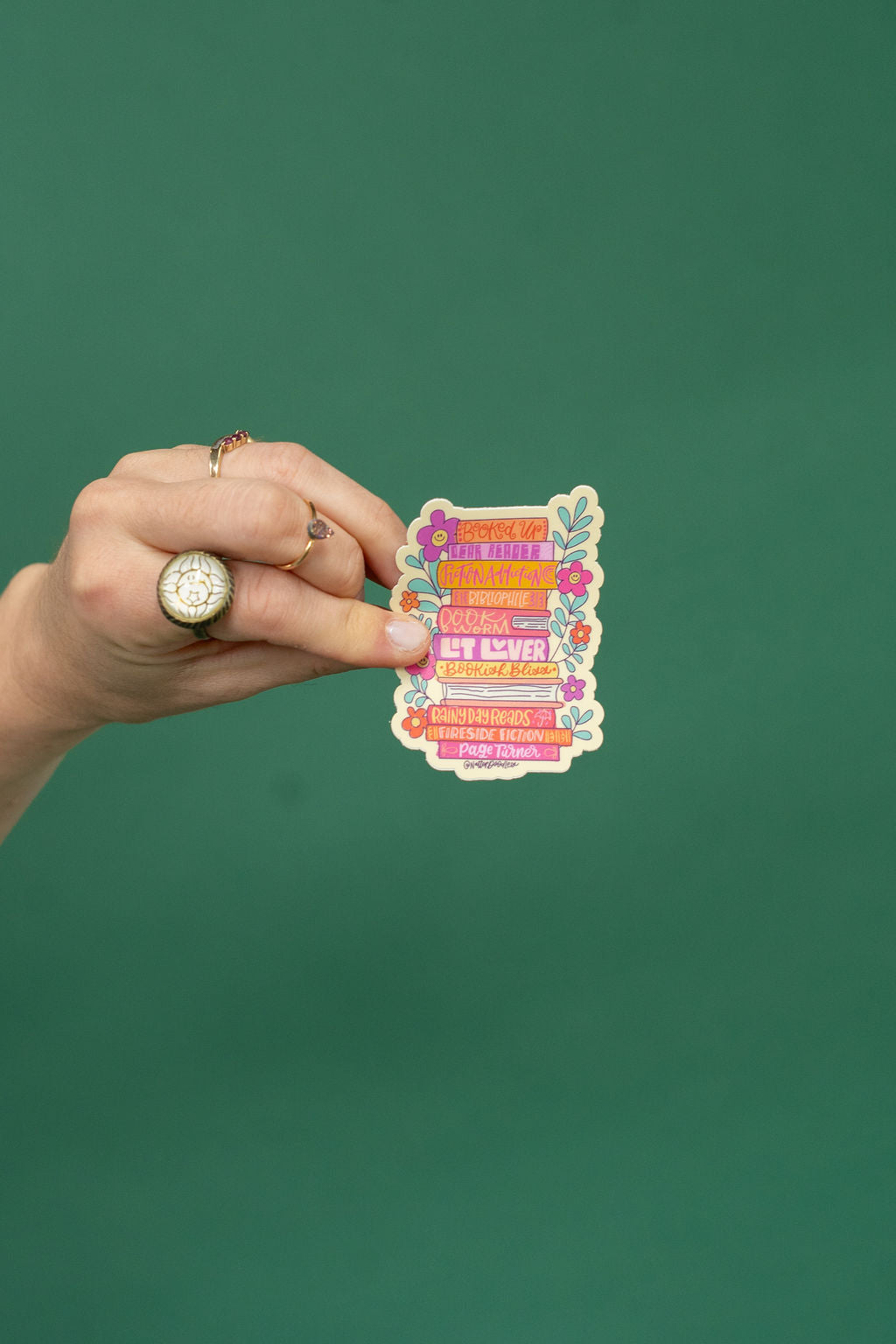 Hand holding a colorful rectangular sticker with text and floral designs against a green background