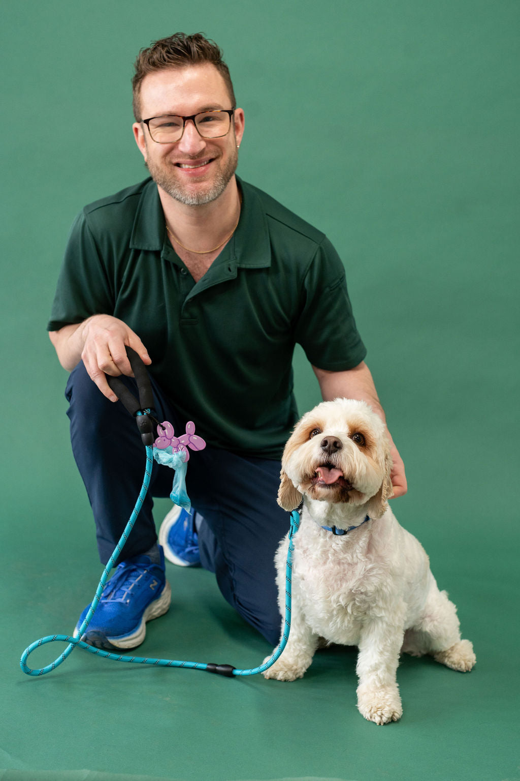 Man kneeling on a green floor with a white dog, holding a blue leash and remote control.