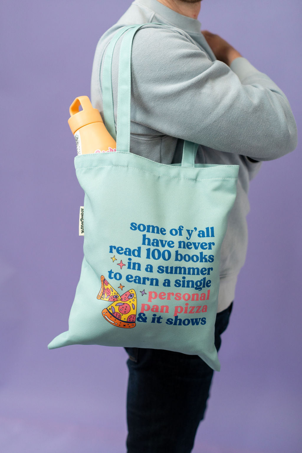 Person holding a light blue tote bag with text and graphics on a purple background