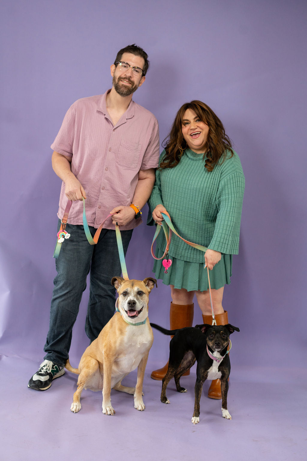 Two people standing with two dogs on leashes against a purple background