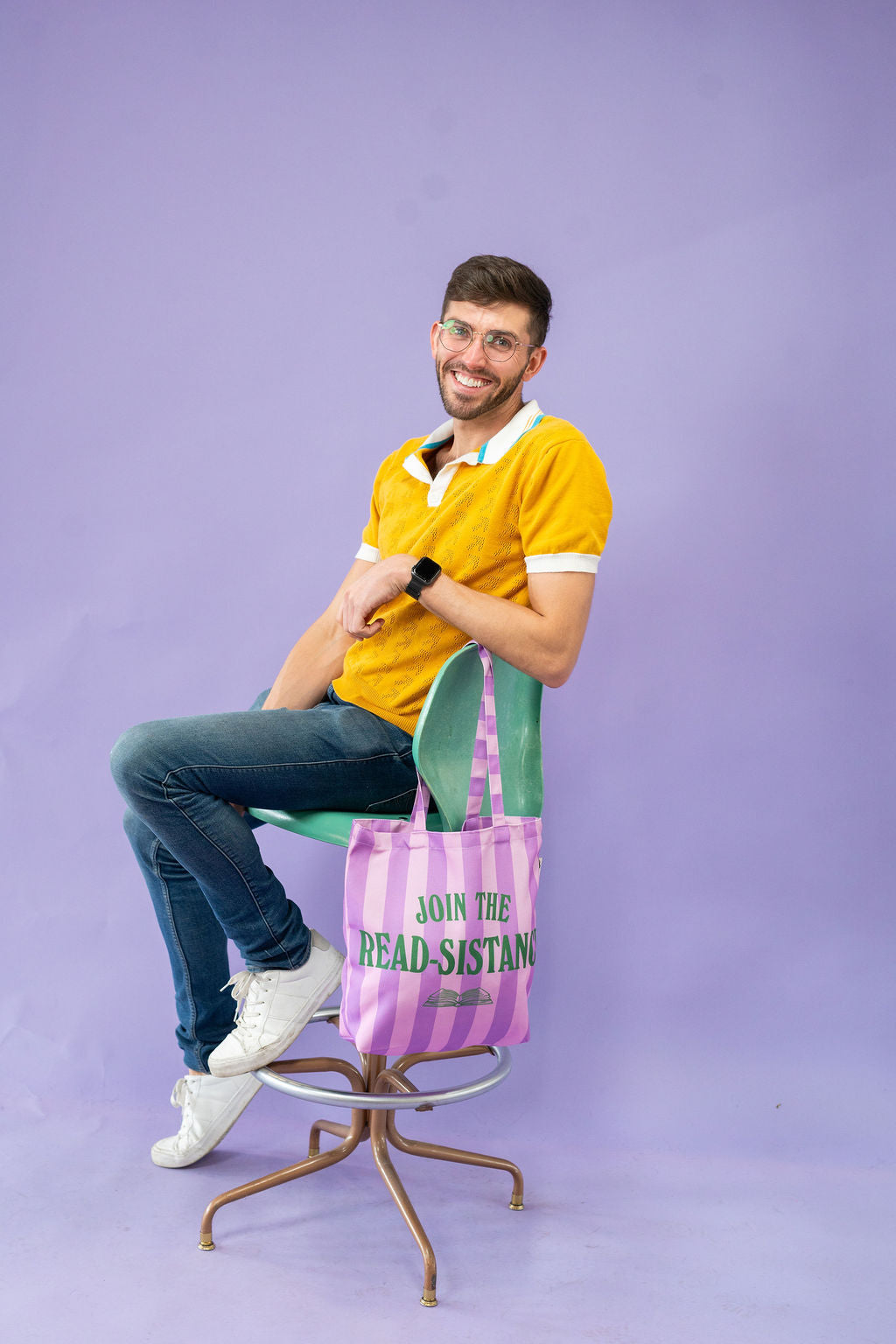 Man sitting on a chair with a striped bag, holding a book, against a purple background