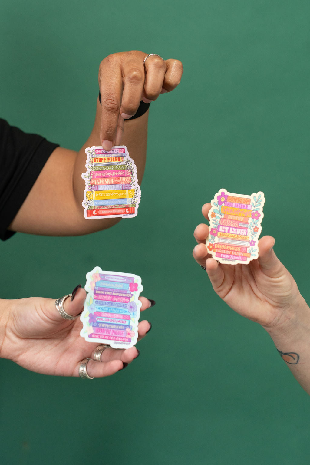 Three hands holding colorful bookish stickers against a green background