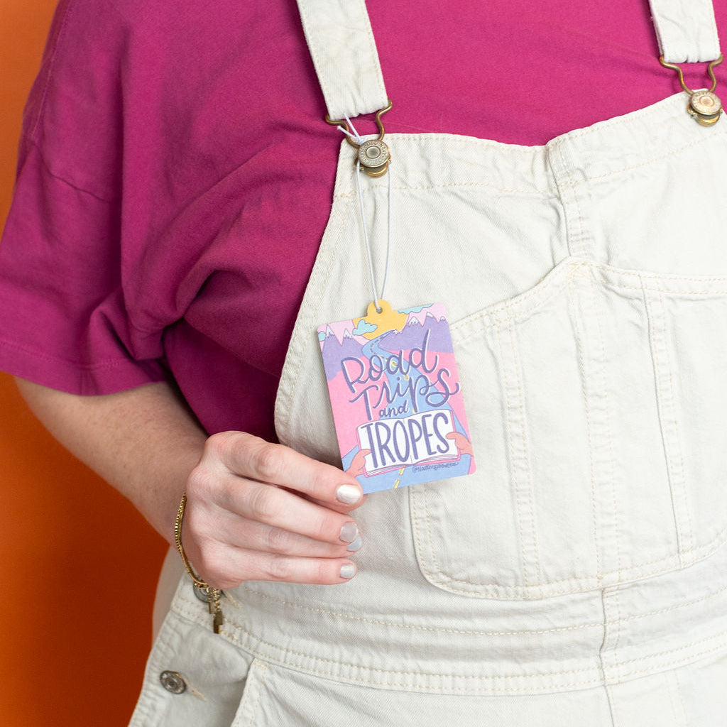 Person wearing a pink shirt and beige overalls holding a colorful card against an orange background