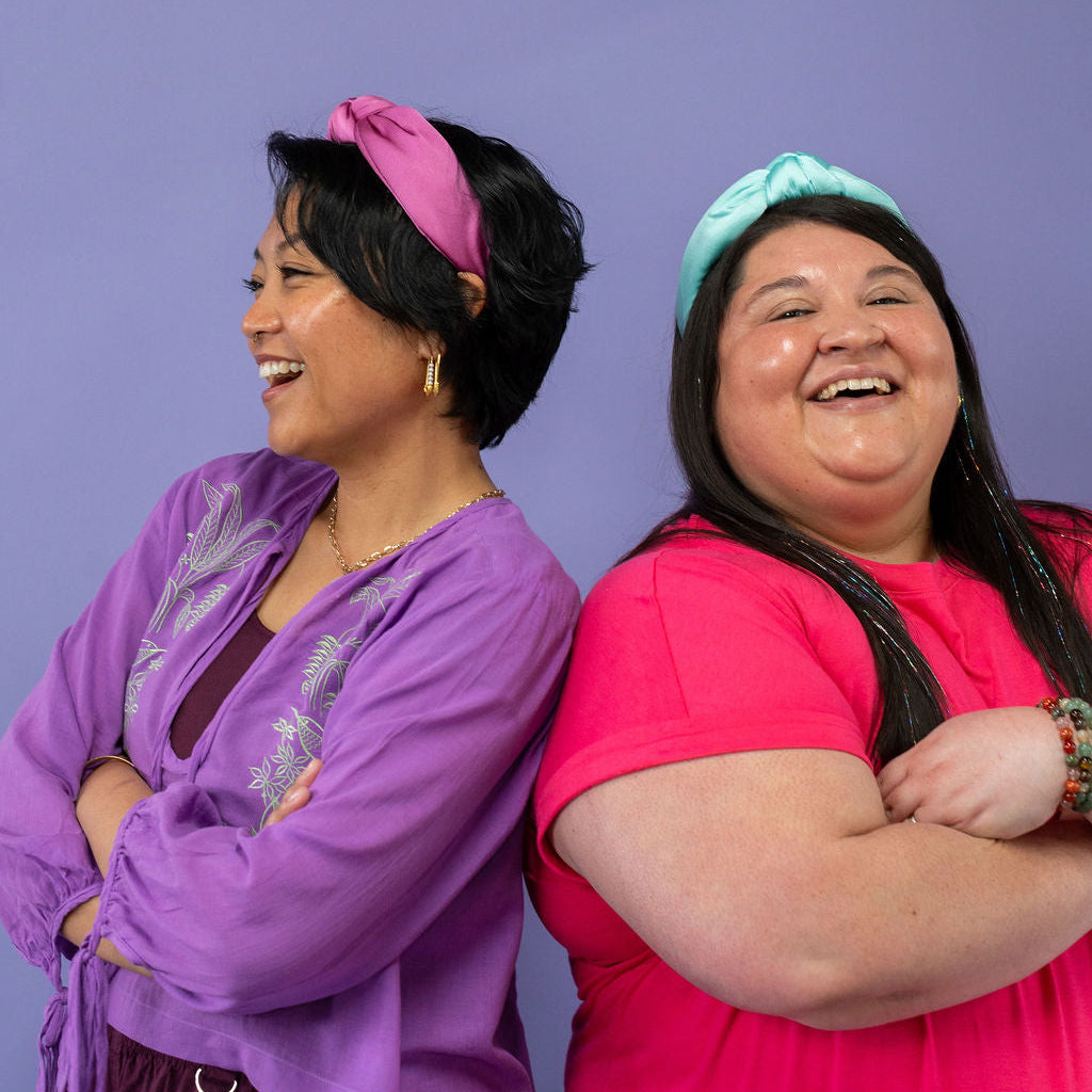 Two women standing back-to-back against a purple background, wearing colorful headbands.