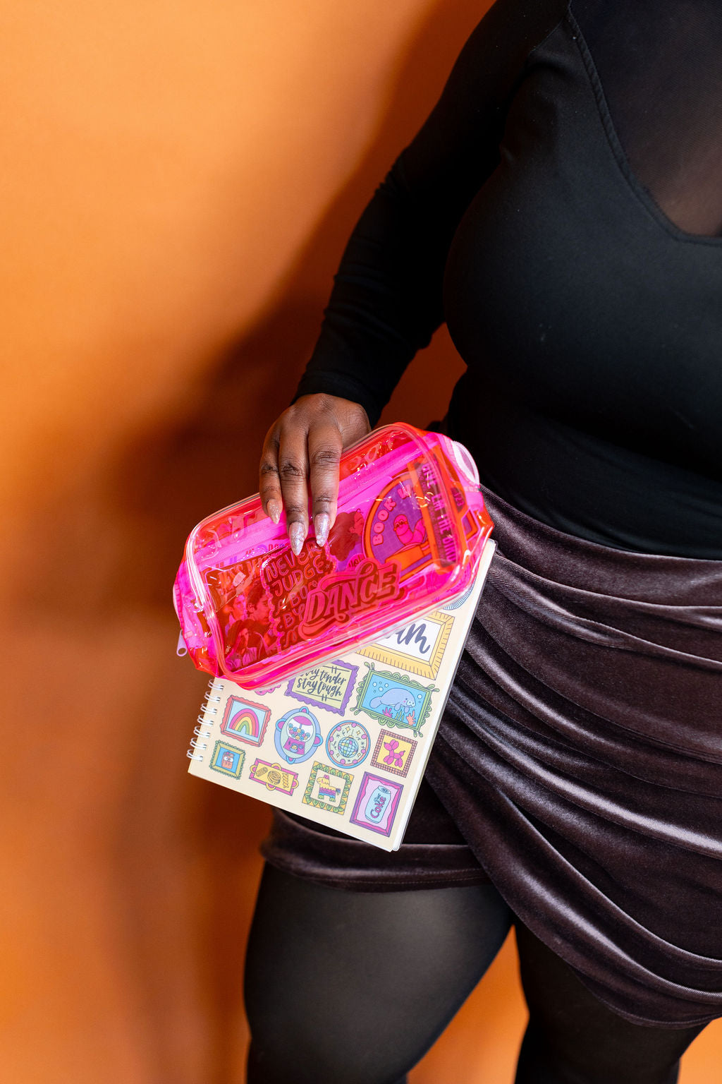 Person holding a pink makeup case and colorful book against an orange background