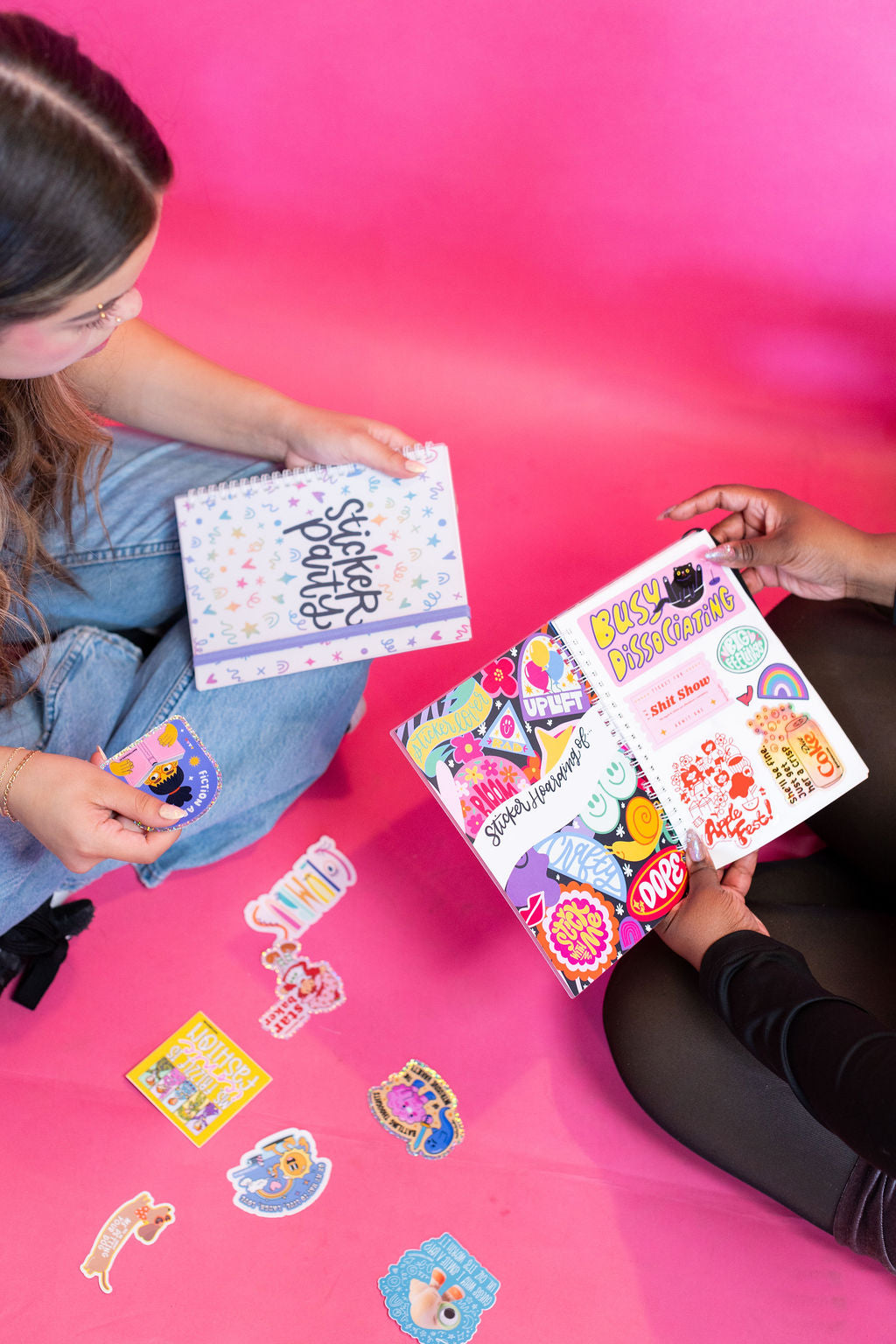 Two people sitting on a pink surface with sticker books and stickers.