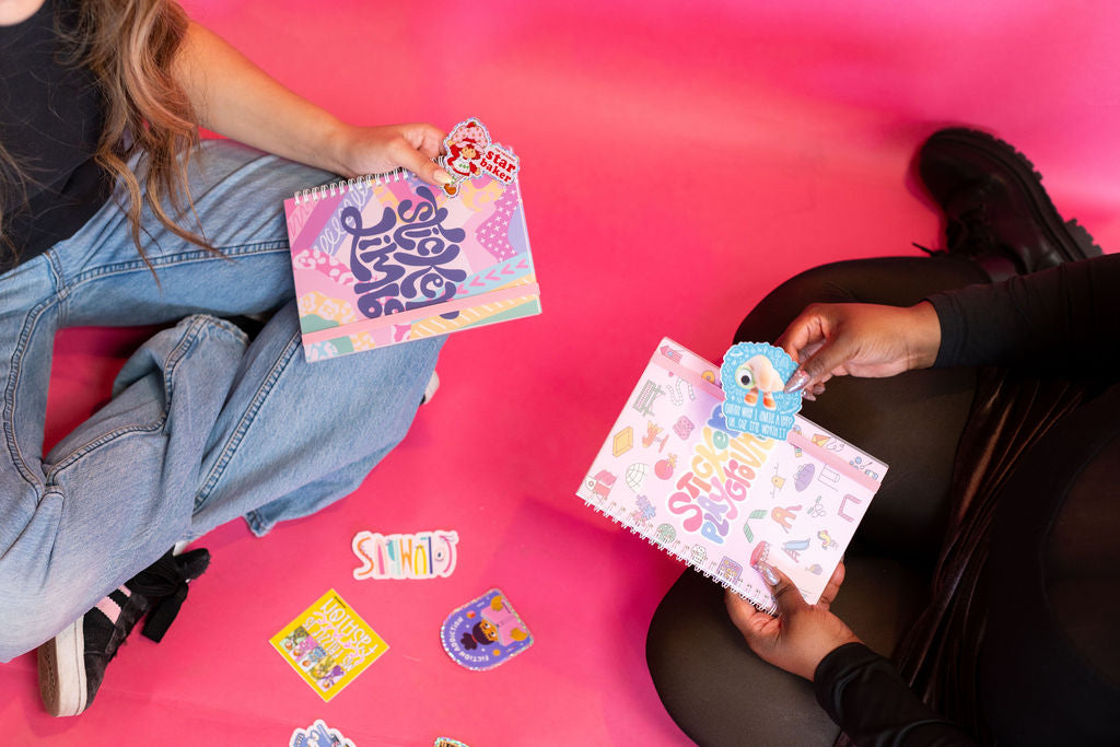 Two people sitting on a pink floor with colorful books and stickers.