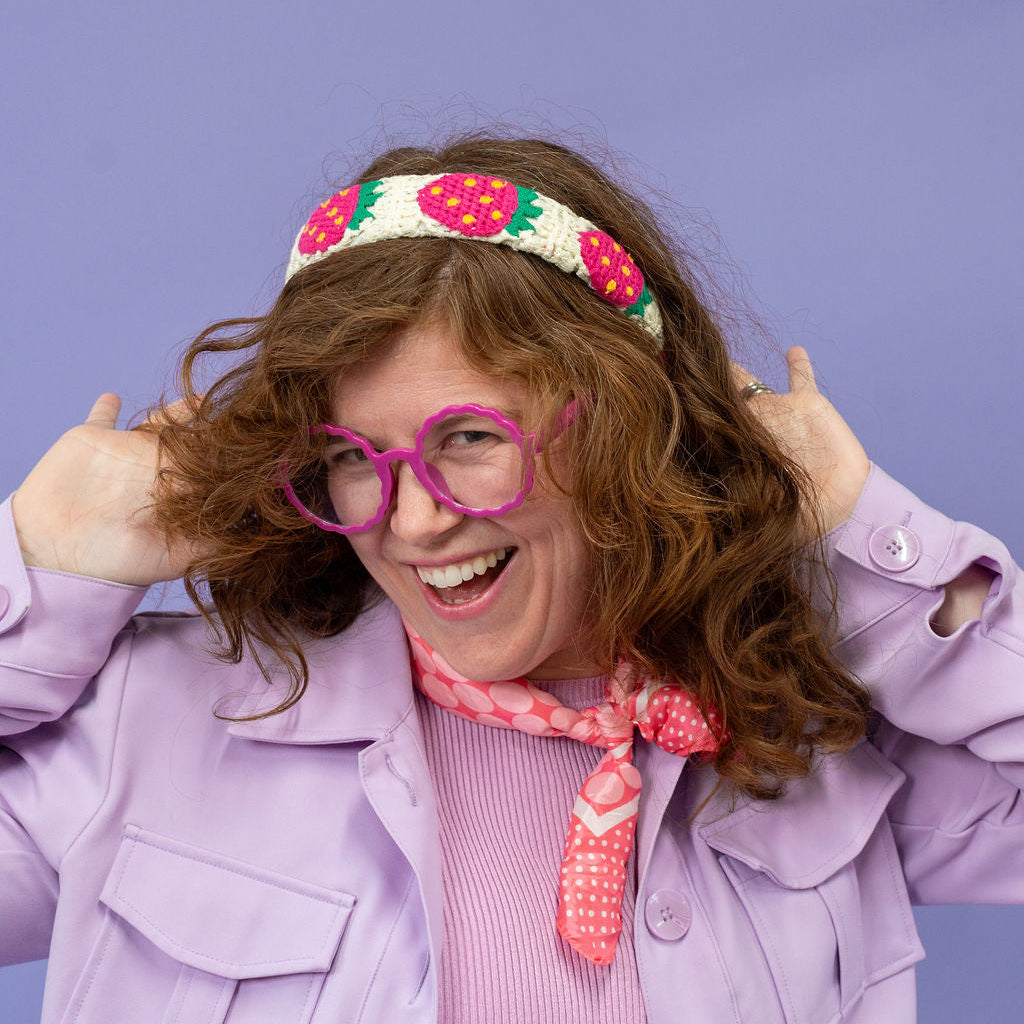 Person wearing a colorful headband, pink glasses, and a purple jacket against a purple background