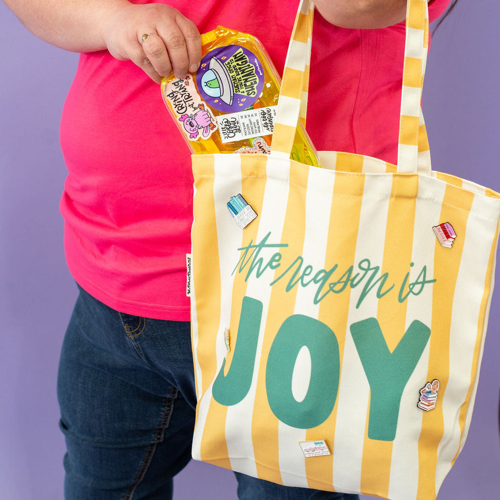Person holding a yellow and white striped tote bag with 'The Reason is Joy' text, against a purple background.
