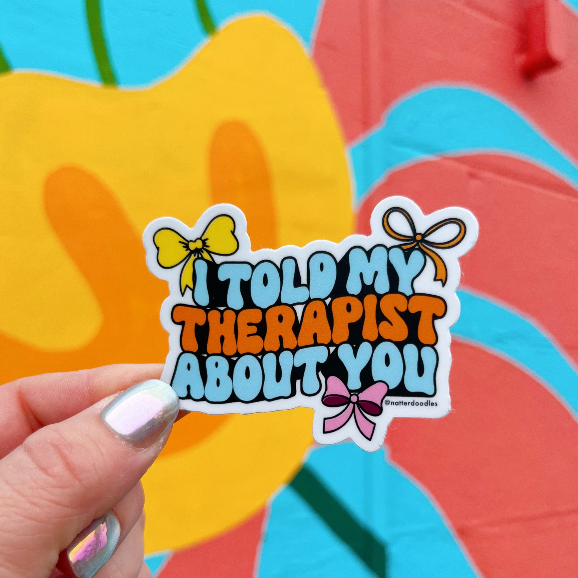 A hand holding a sticker with the text 'I Told My Therapist About You' written in a hand-lettered style, accompanied by colorful graphic elements such as bows.