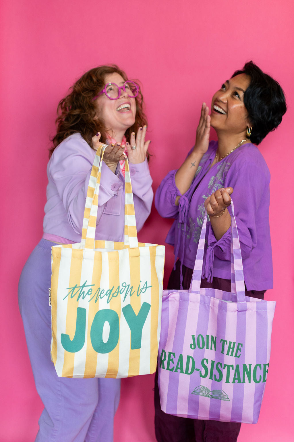 Two women holding tote bags with motivational phrases against a pink background