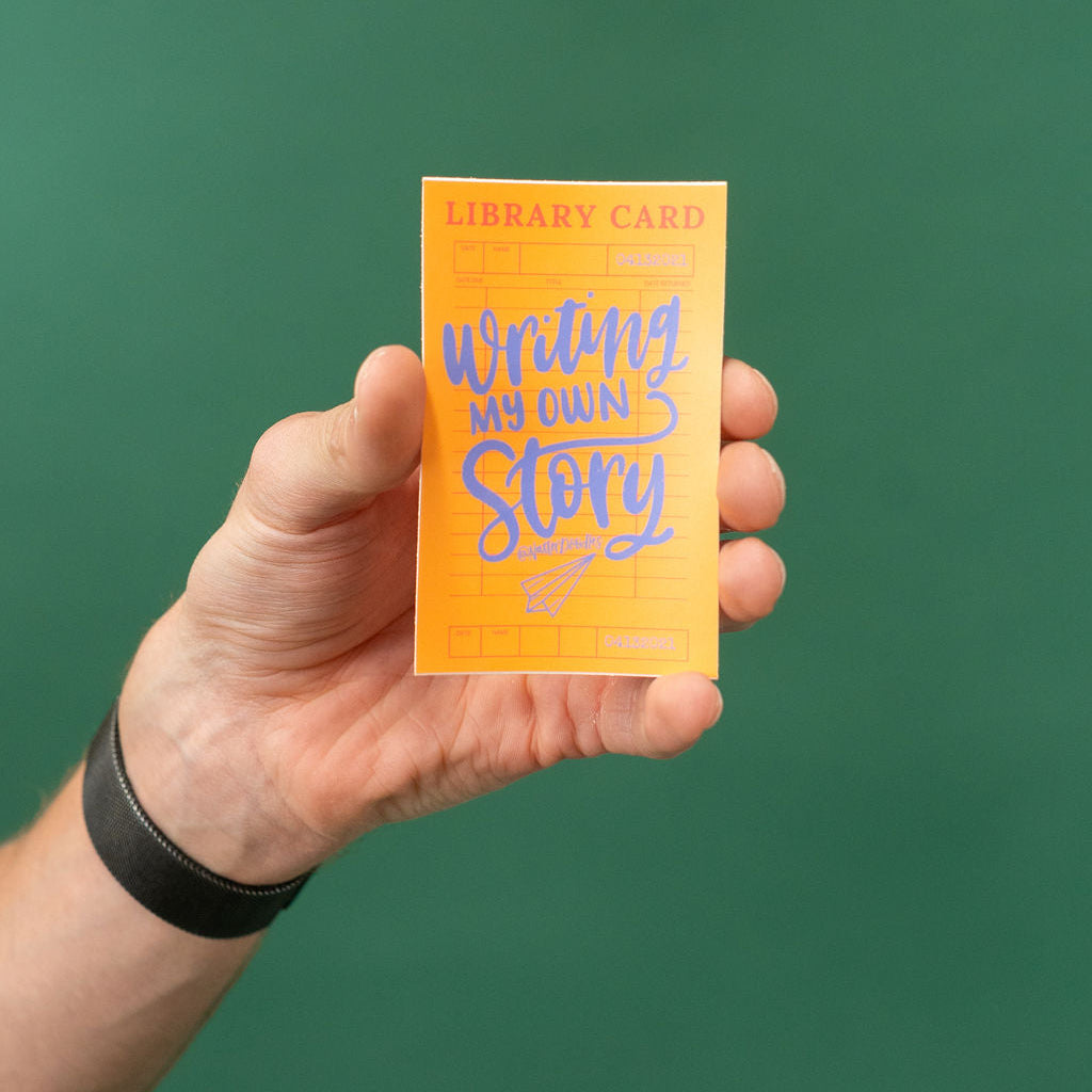Hand holding a yellow library card with 'Writing My Own Story' text against a green background