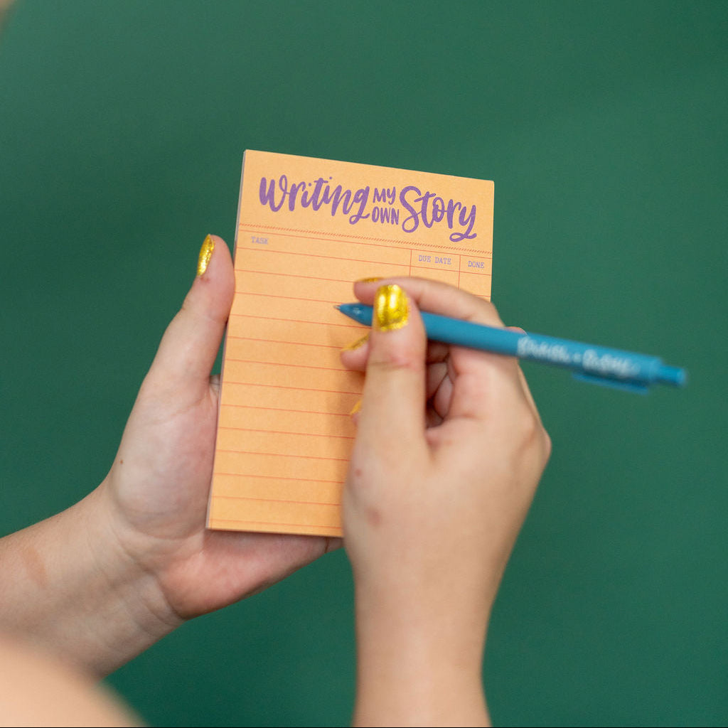 Hand holding a small notepad with 'Writing My Story' text against a green background