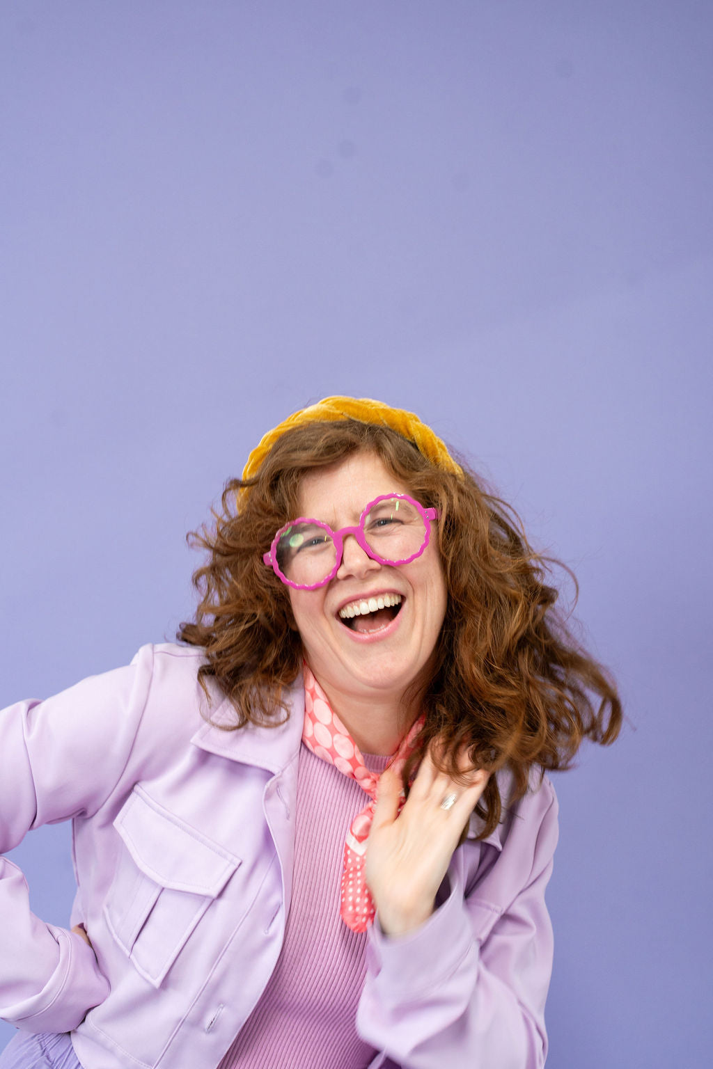 Person wearing a purple jacket, pink glasses, and a yellow hat against a purple background