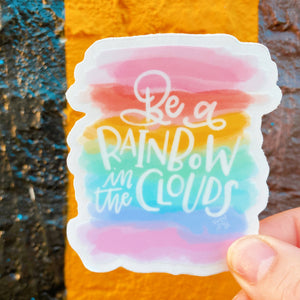 Die cut sticker features a watercolor rainbow background and the phrase "be a rainbow in the clouds"